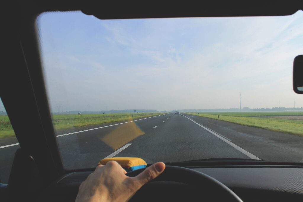 View from a car through the windshield, driving down an empty highway with clear skies and green fields.